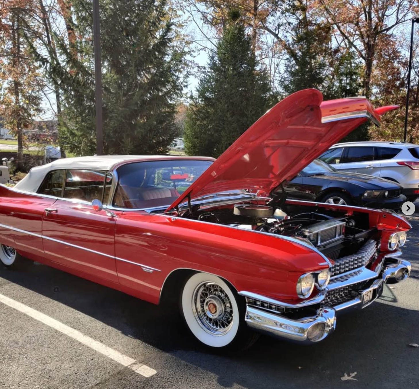 A bright red vintage Cadillac convertible with its hood raised, parked in an outdoor lot on a sunny day.