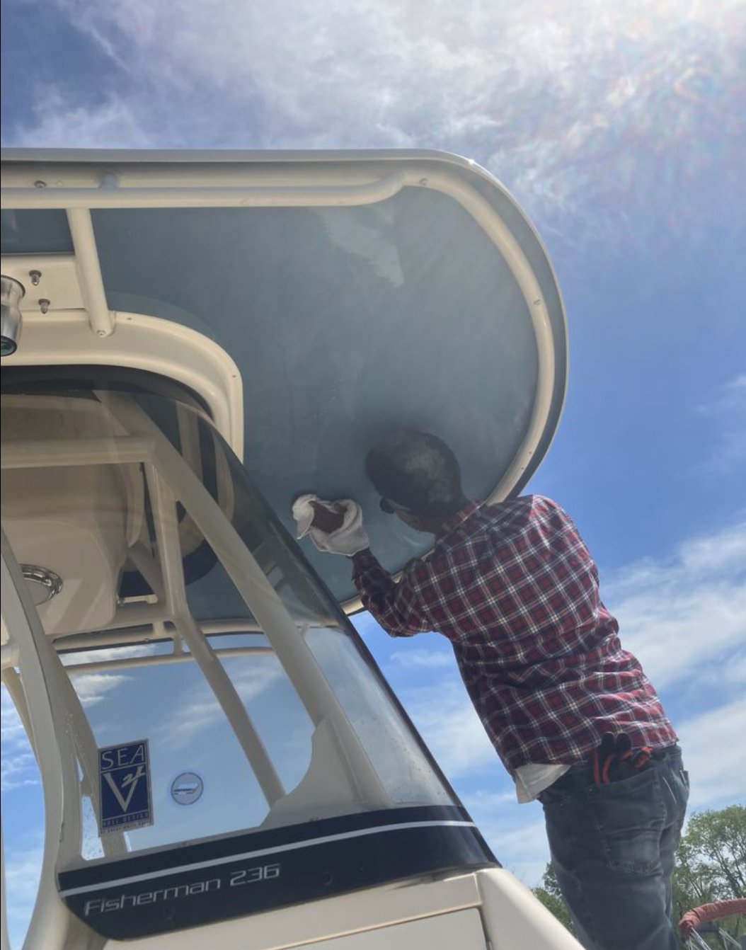 A person in a plaid shirt wiping the underside of a boat's hardtop roof against a blue sky.