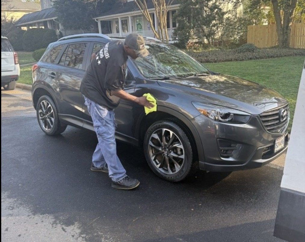 A person in a black shirt and jeans wipes the side of a dark gray SUV parked on a paved driveway.
