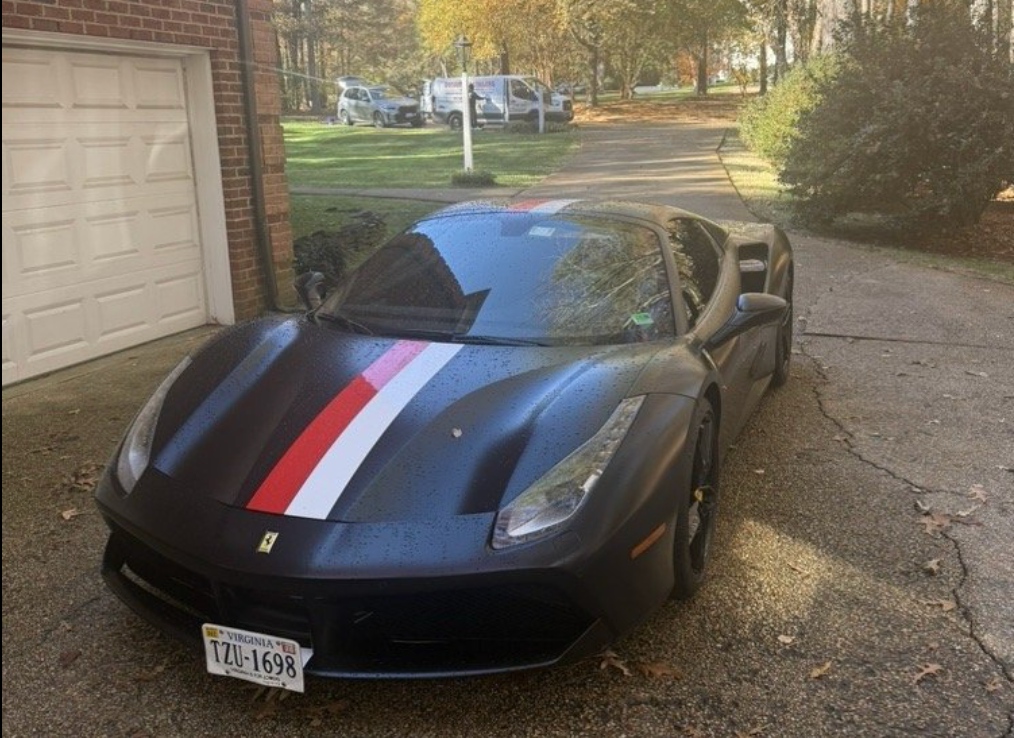 A black Ferrari sports car with a red and white racing stripe parked on a residential concrete driveway.
