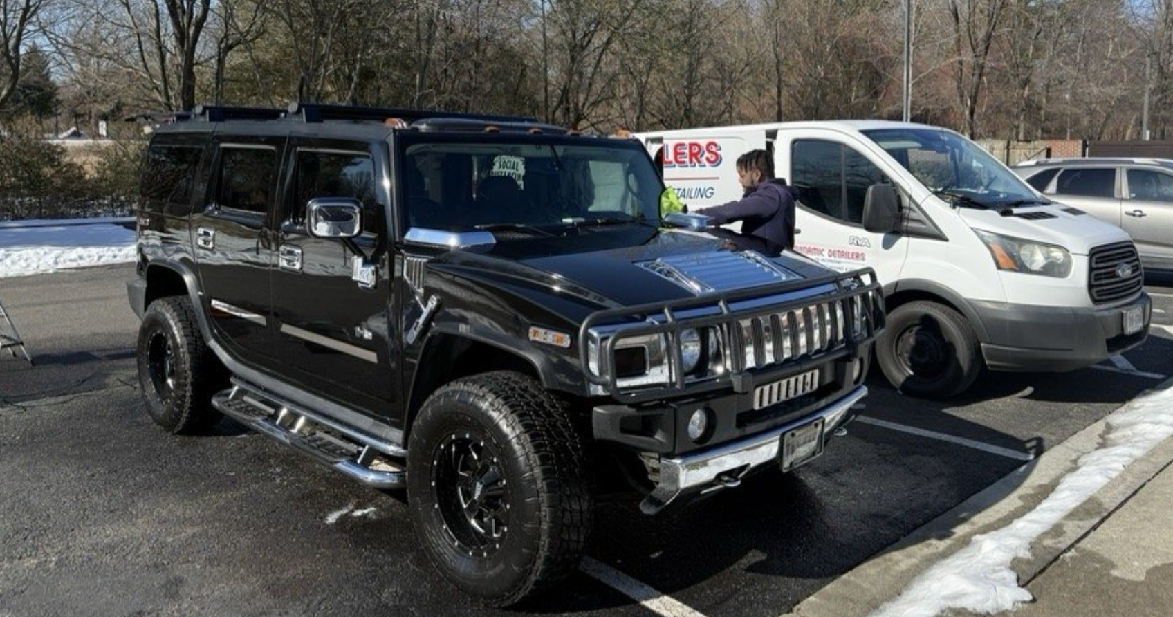 A black Hummer SUV parked next to a white service van in a paved lot with patches of snow on the ground.