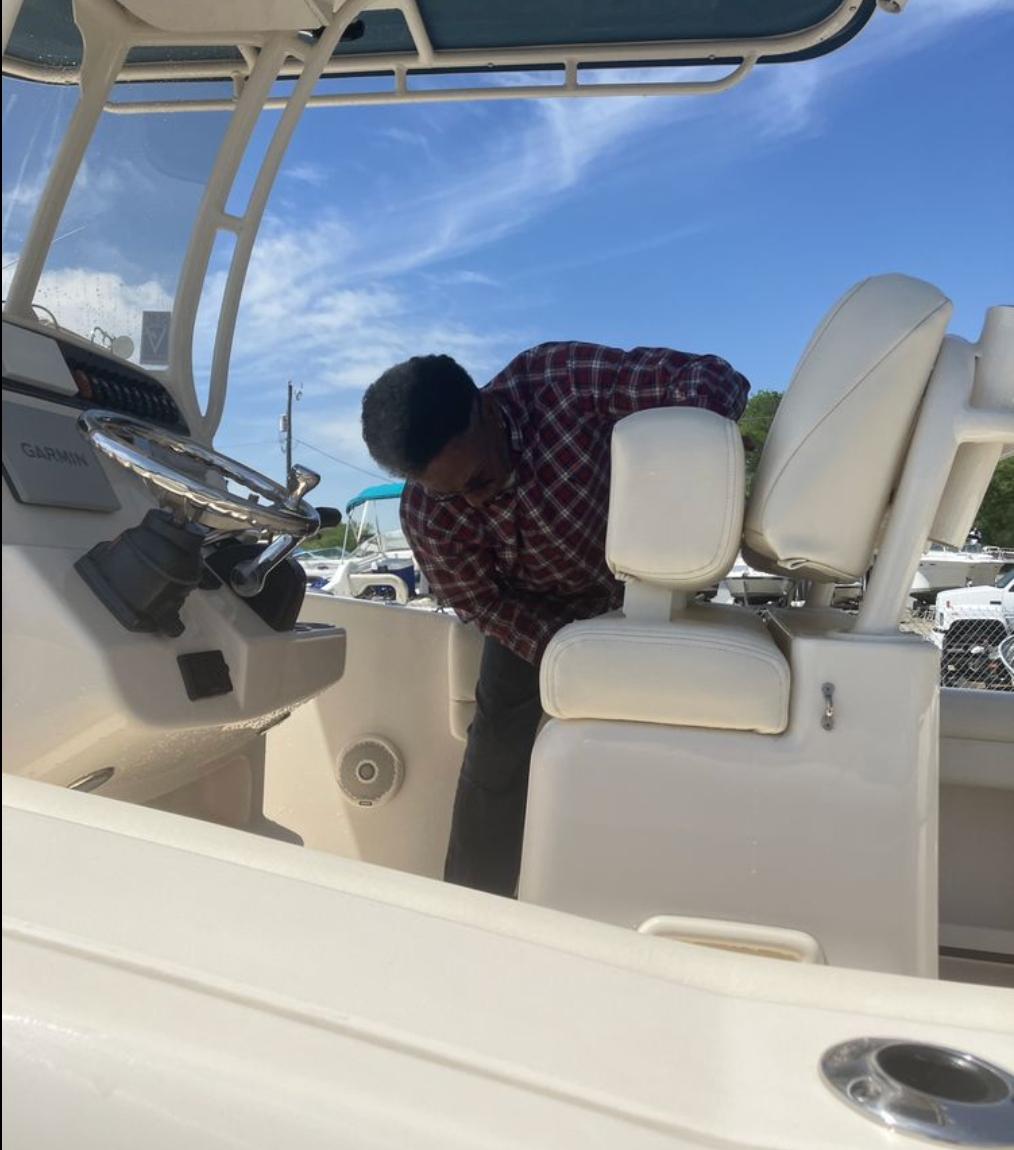 A person in a plaid shirt inspects a boat's helm area, standing near the white seats under a sun-covered roof.