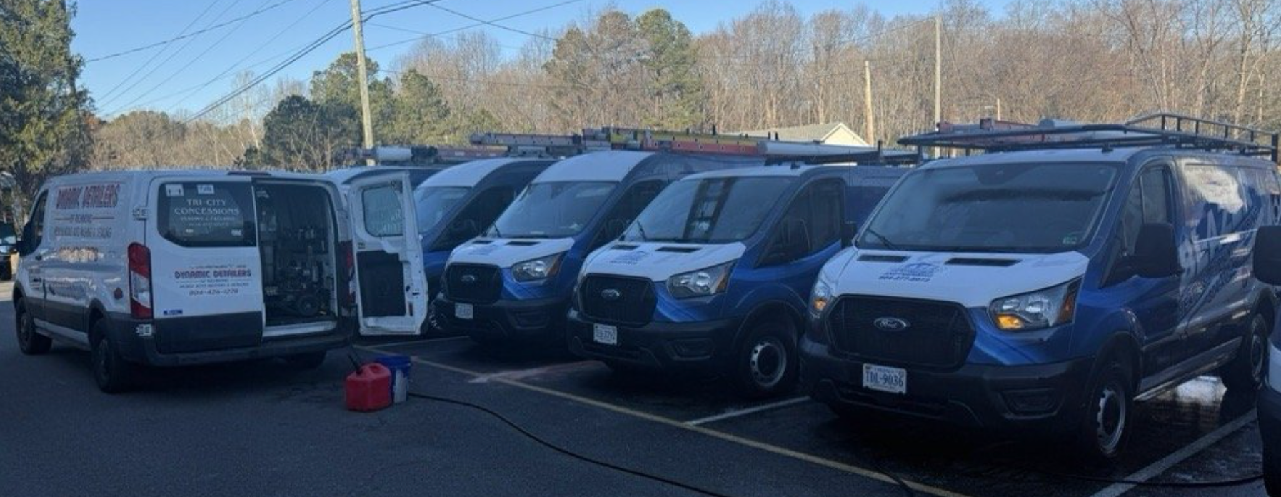 A row of parked white and blue service vans with roof racks, one with its rear doors open, in a sunny outdoor lot.
