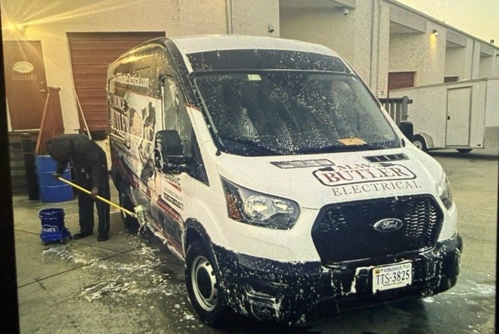 A person washing a white Butler Electrical work van with a long-handled brush outside a commercial building.