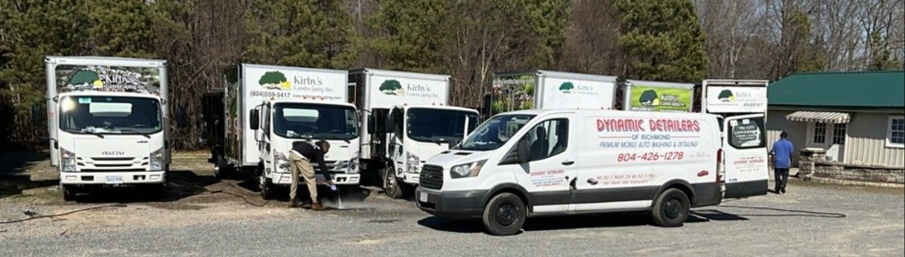 A row of service trucks and a van parked in a gravel lot in front of a small building with a green roof.