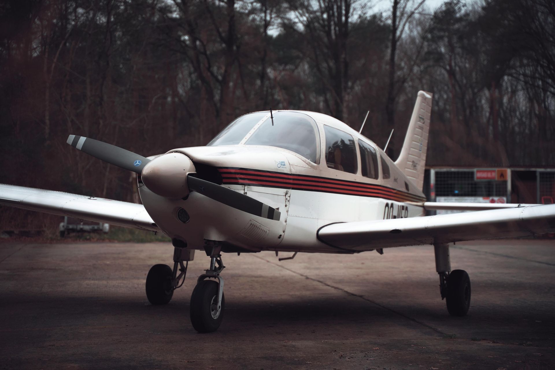 A white single-engine propeller plane with red stripes parked on a paved runway in front of trees.