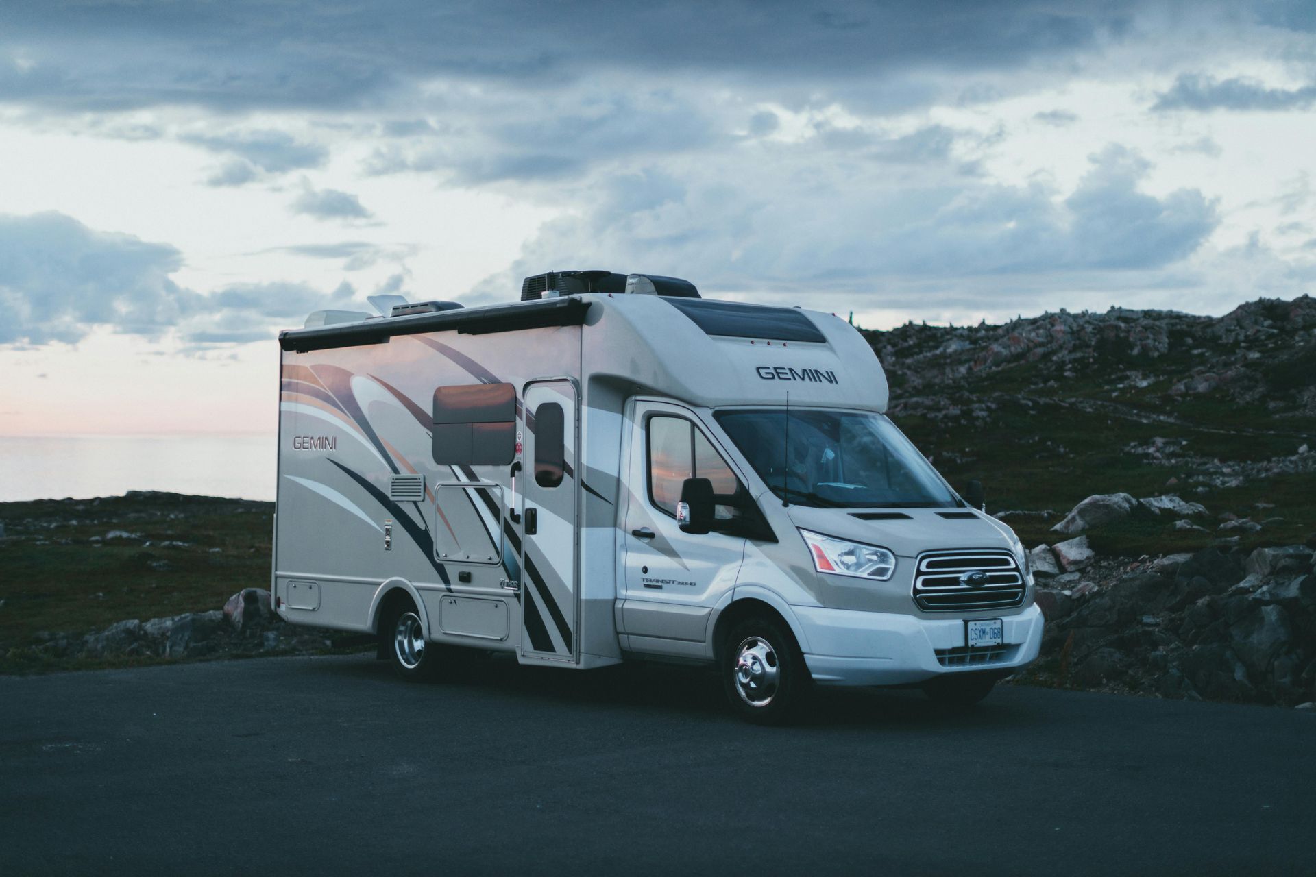 A modern beige and white recreational vehicle parked on a paved road against a dramatic, cloudy sunset sky.