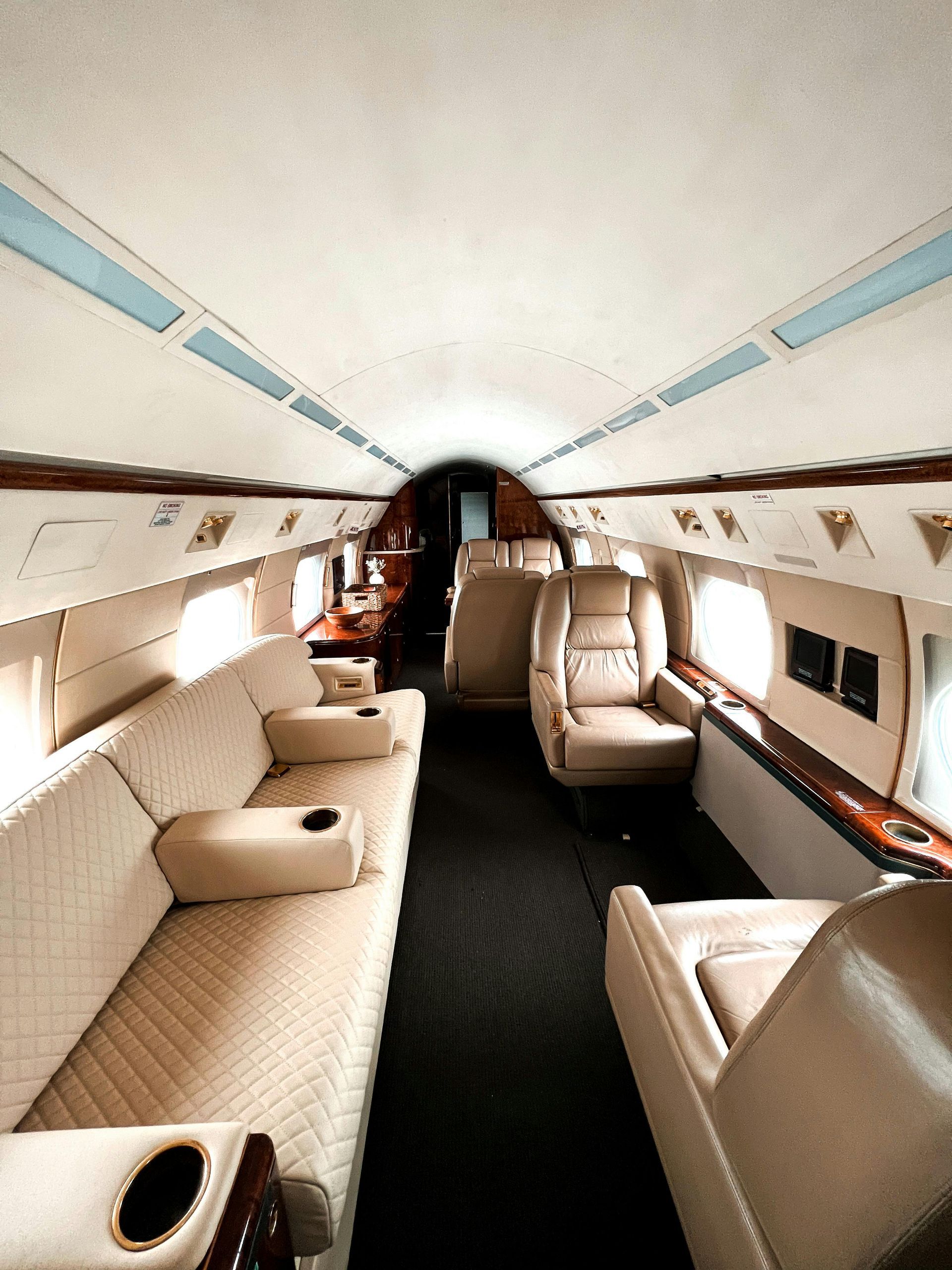 The interior of a private jet cabin featuring a long cream-colored sofa, leather seats, and dark carpet.