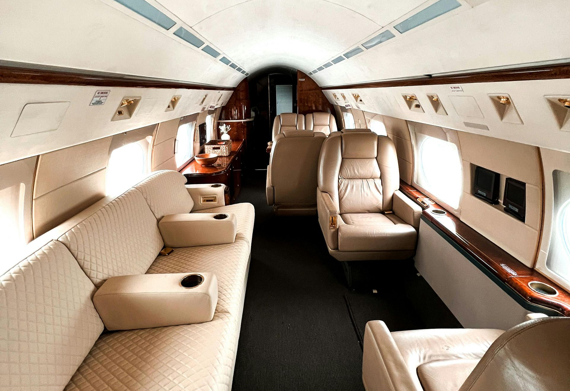 The interior cabin of a private jet with cream-colored leather seats, a long sofa, and wood accents.