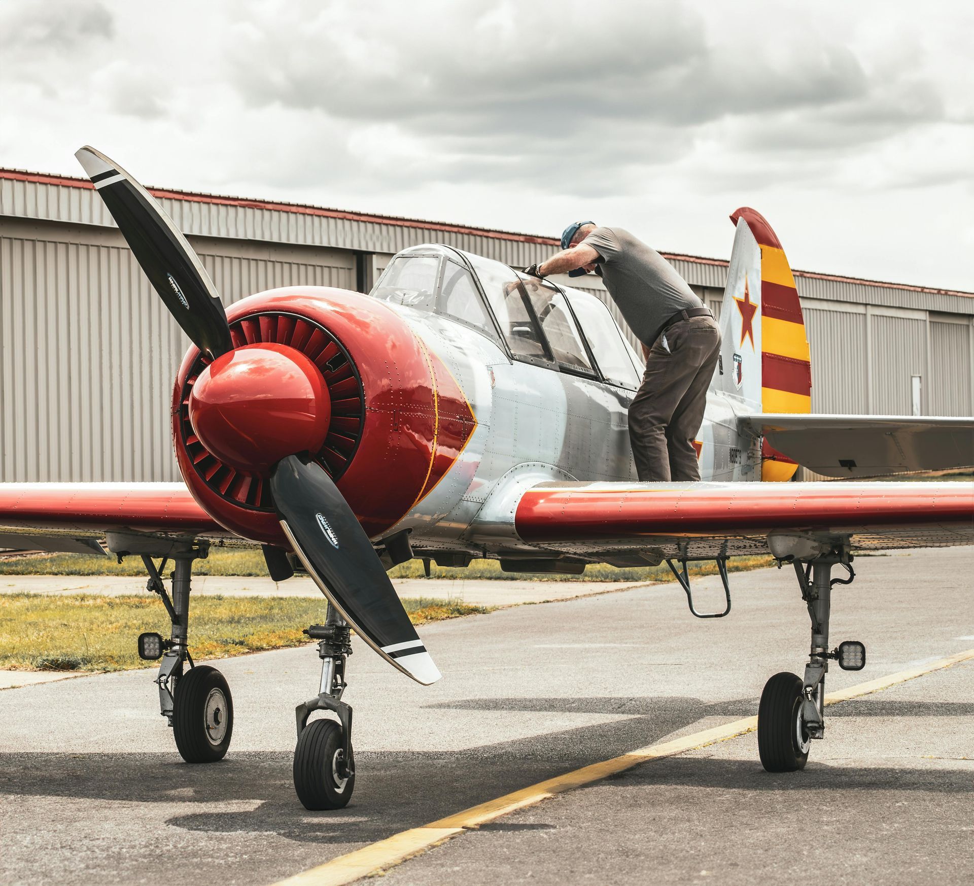 A person stands on the wing of a red and white propeller plane, inspecting or cleaning the cockpit canopy.