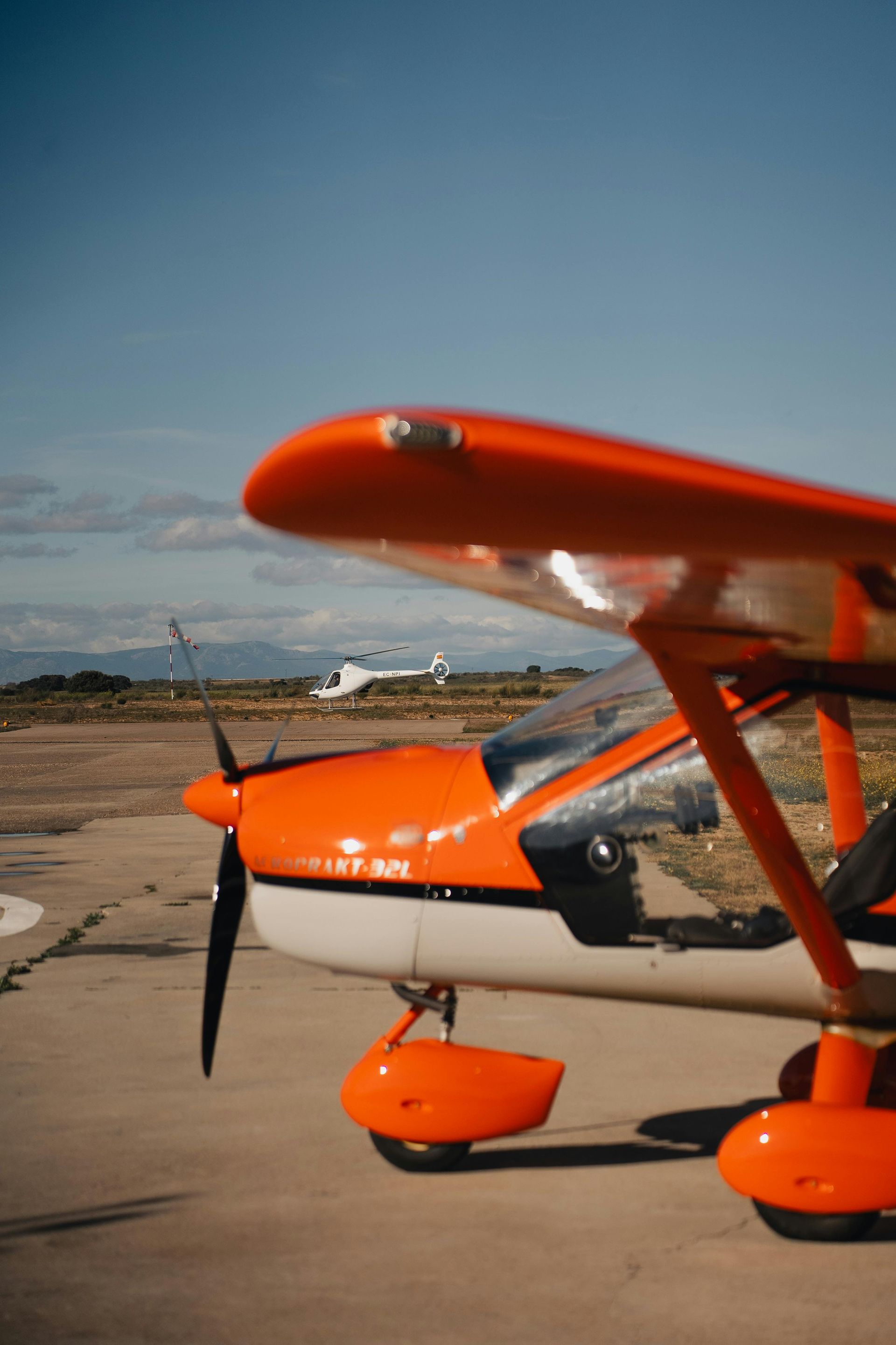 A bright orange and white light aircraft parked on an airfield with a small helicopter visible in the distance.