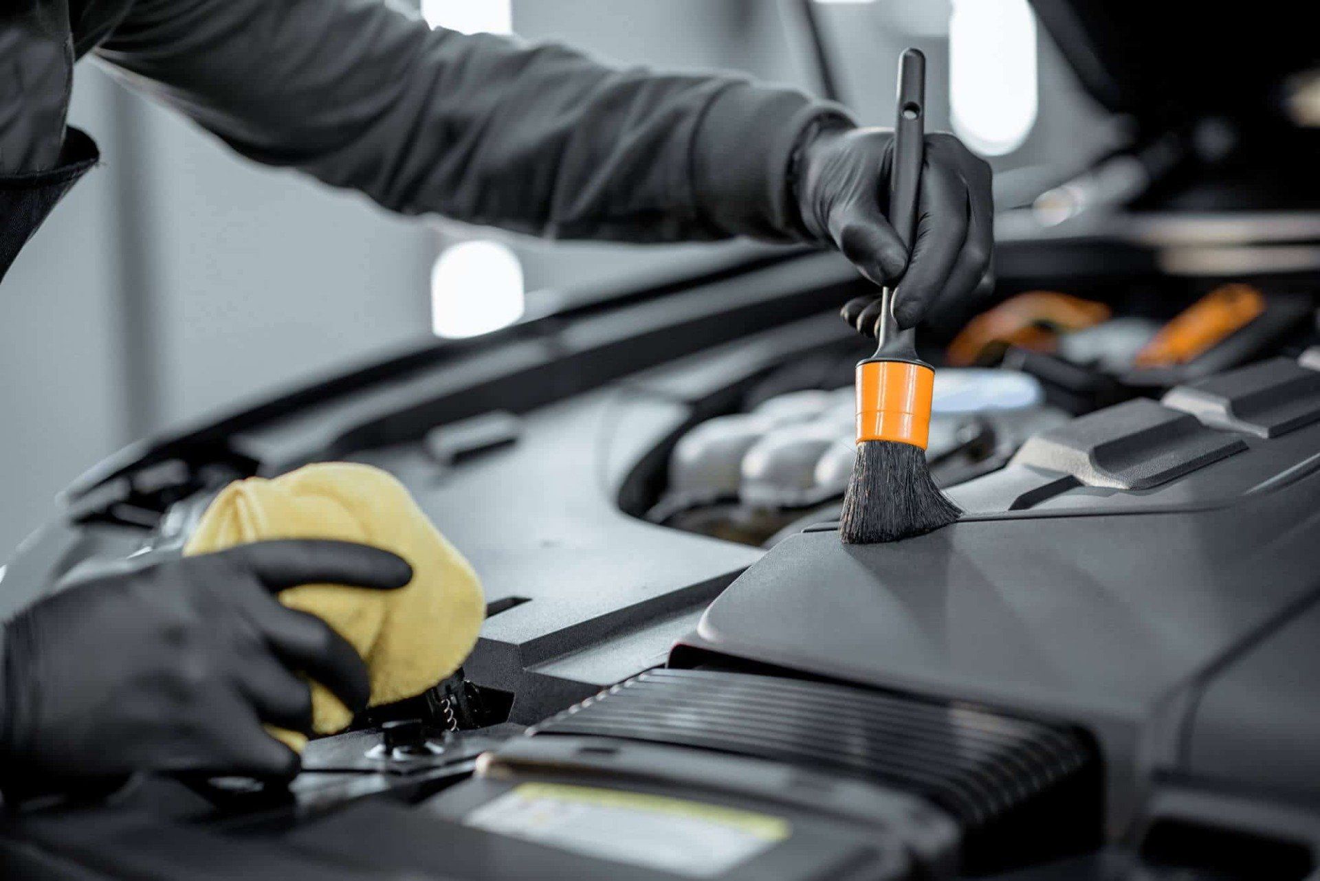 Mechanic wearing black gloves uses a small brush to clean the engine bay of a car.