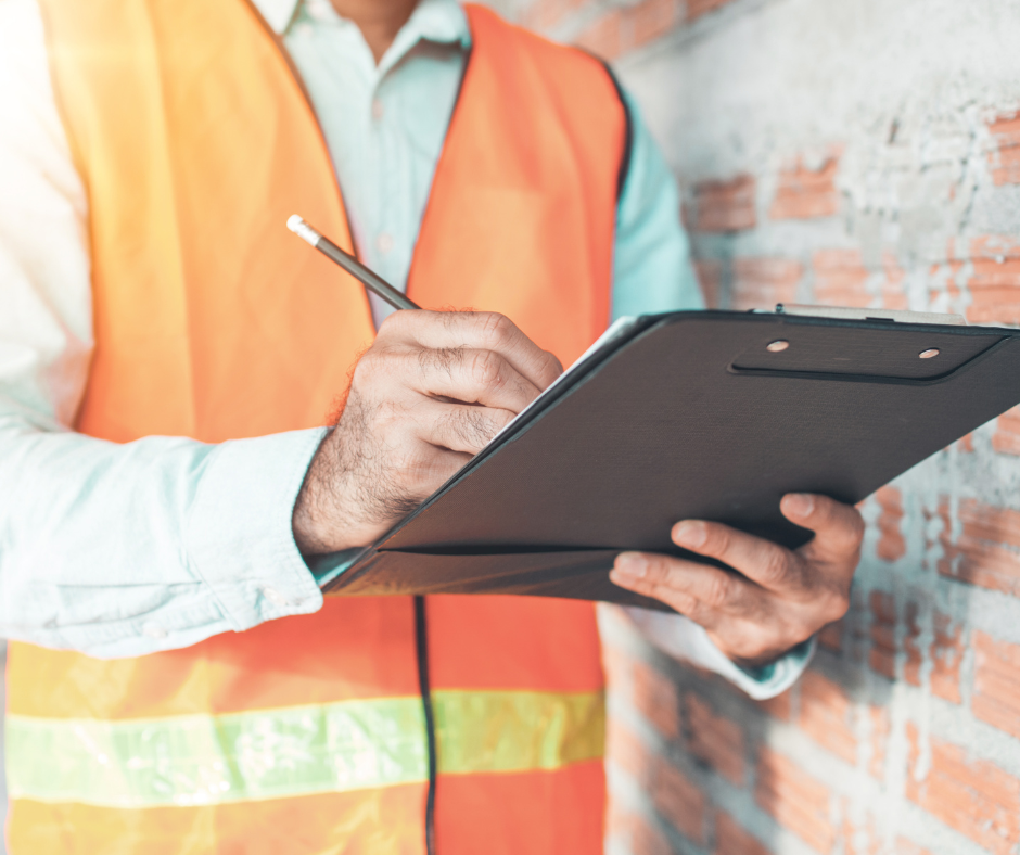 Construction worker in orange safety vest writing on a clipboard beside a brick wall