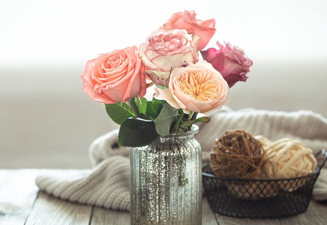 Bouquet of pink and peach roses in a glass vase, next to yarn balls and a sweater.