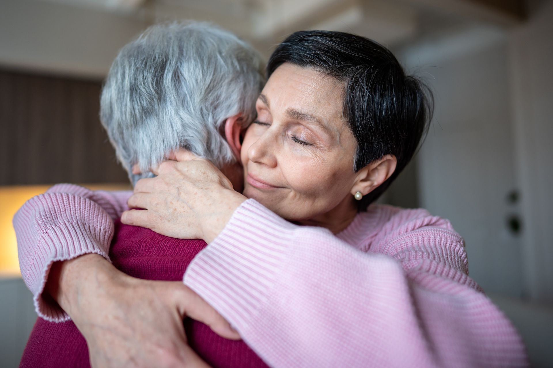 Woman embracing another with eyes closed, pink sweater, soft lighting.