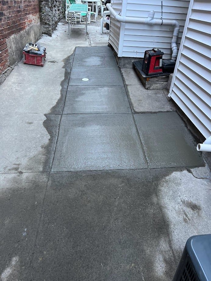 A freshly poured concrete path sits between an exterior brick wall and the white siding of a house.