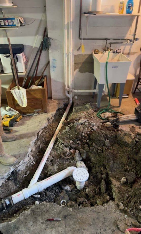 A basement utility room with a trench cut into the concrete floor, showing newly installed white PVC plumbing and a drain.