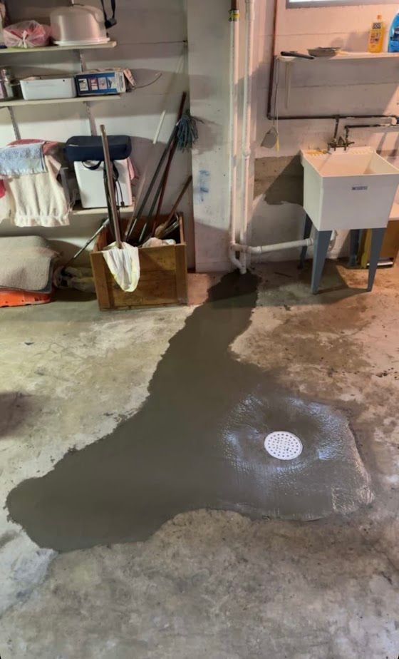A basement floor with a fresh patch of wet concrete around a circular floor drain, near a utility sink and shelving.