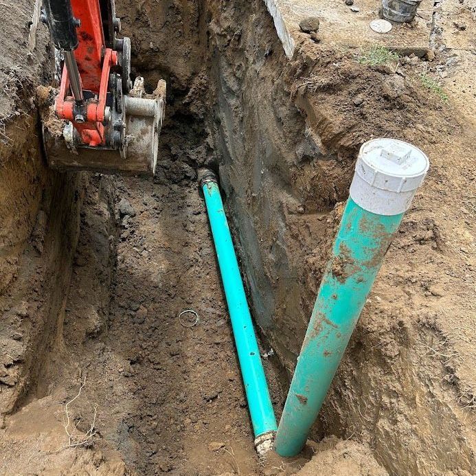 An excavator bucket is positioned above a green sewer pipe in a trench, featuring a vertical cleanout access pipe.