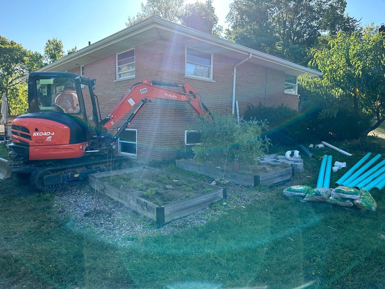 An orange mini excavator positioned near the brick corner of a residential house with landscaping materials on the lawn.