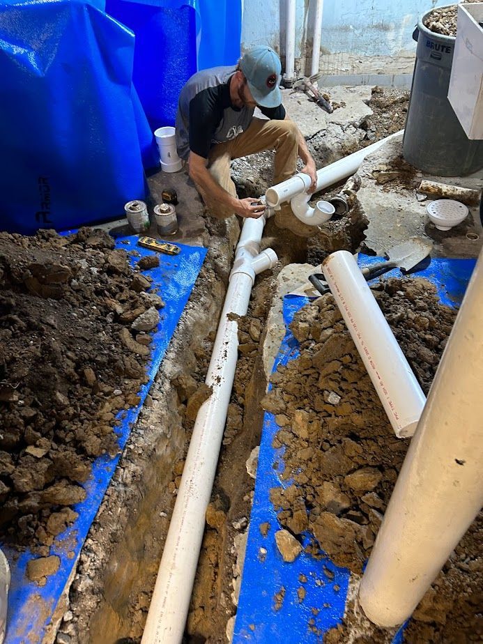 A person wearing a grey shirt and cap works on installing white PVC plumbing pipes in a dirt trench lined with blue plastic.