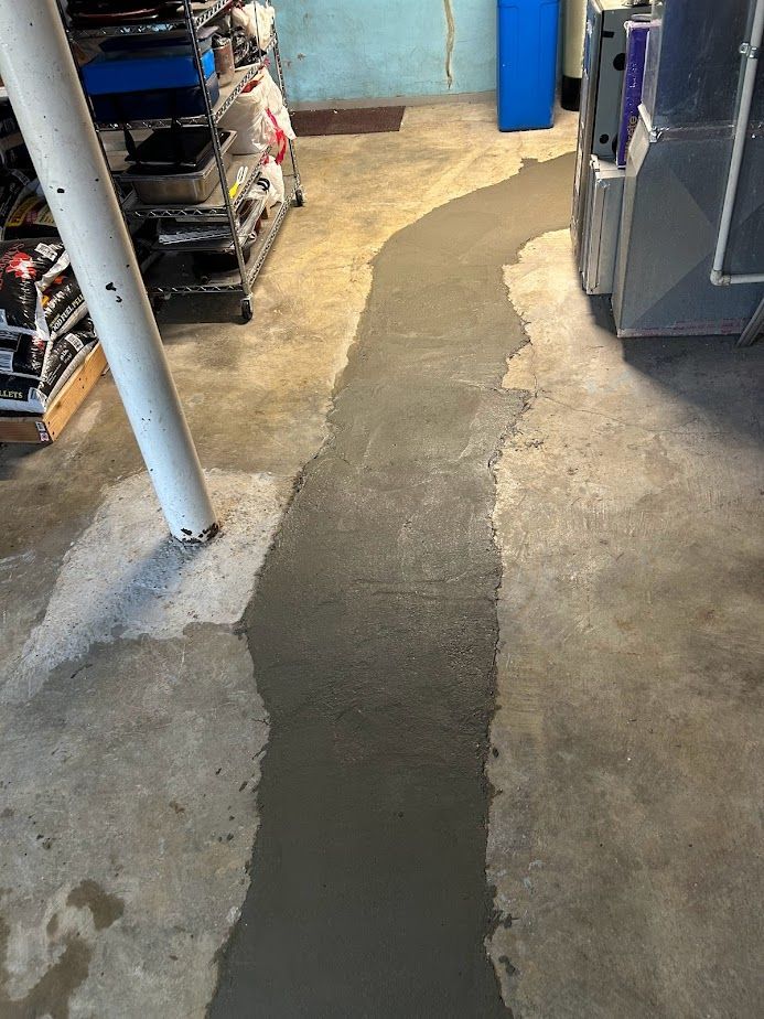 Fresh concrete patch filling a narrow trench across a concrete basement floor near a metal pole and storage shelving.