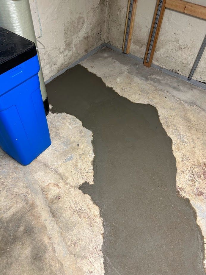 A patch of fresh, gray concrete fills a trench in a concrete basement floor next to a blue plastic container.