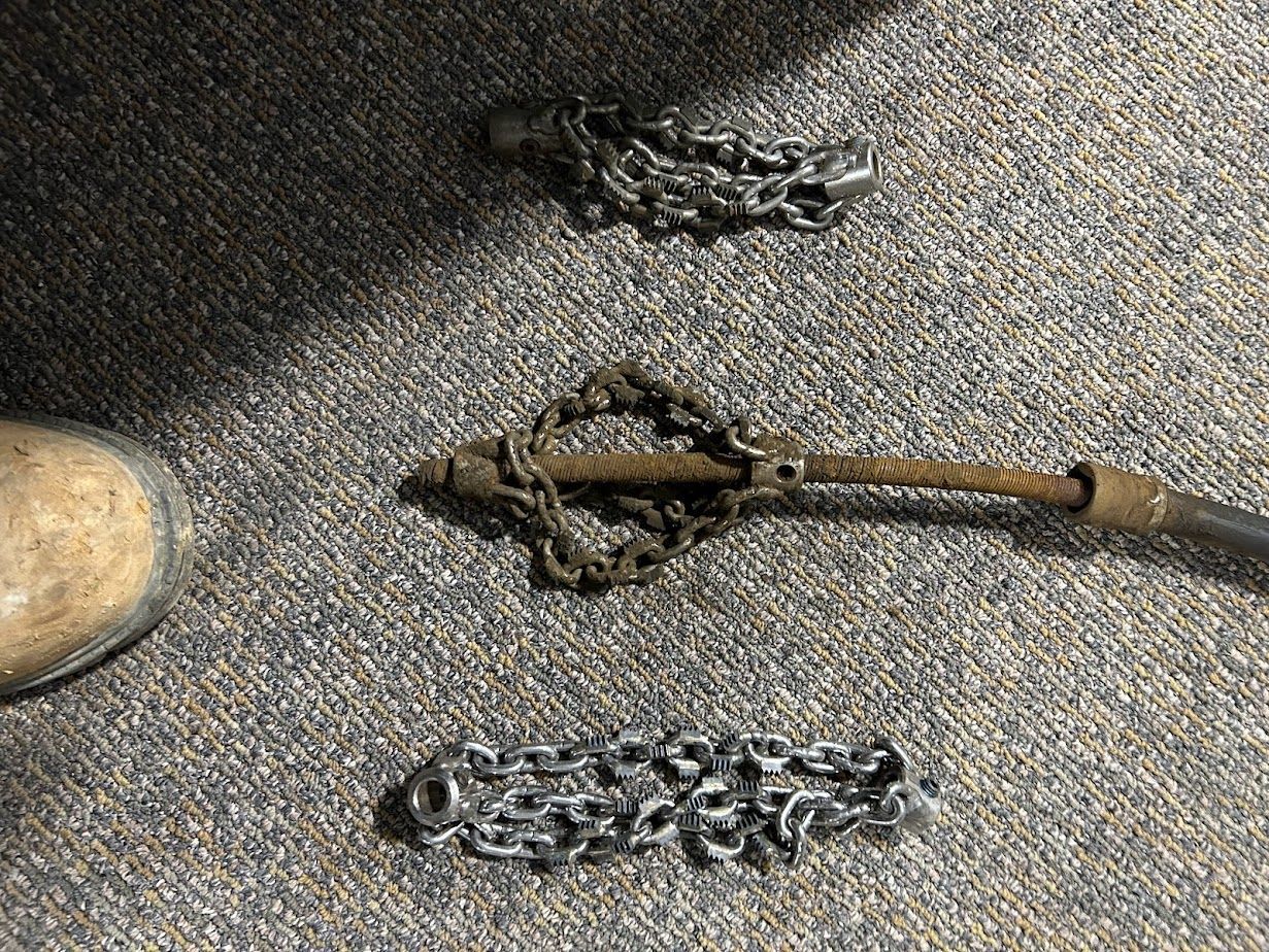 Three lengths of industrial chain and a pipe-cleaning tool with rotating chains rest on a grey carpet next to a boot.