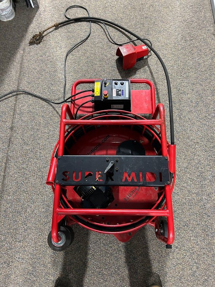 A red Super Mini drain cleaning machine with a foot pedal control on a carpeted floor.
