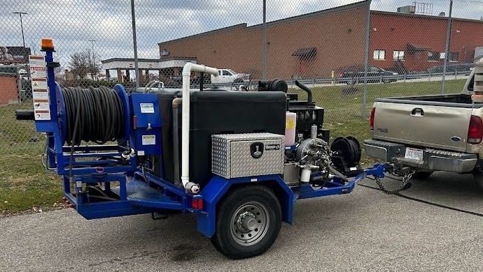 A blue trailer-mounted jetter system with a black water tank and a hose reel, attached to a pickup truck in a parking lot.