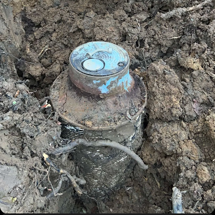 A metal water meter housing with a blue lid, partially excavated from wet, dark soil.
