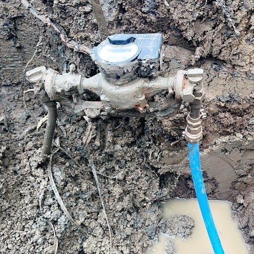 A water meter connected to a blue pipe, exposed in a muddy, excavated trench.