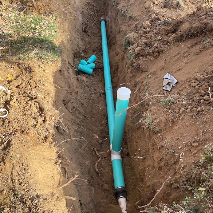 A green PVC pipe with a vertical branch and white cap installed in an open dirt construction trench.