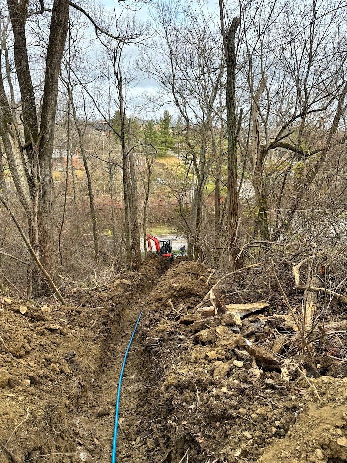 A blue utility pipe lies in a freshly dug dirt trench through a wooded area, leading toward a distant piece of machinery.