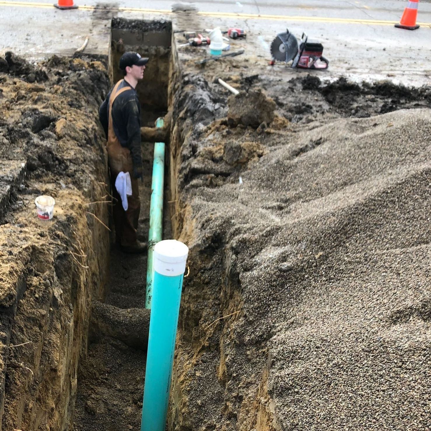 A person in work coveralls stands in a deep dirt trench next to a turquoise pipe, with a stone pile and saw nearby.