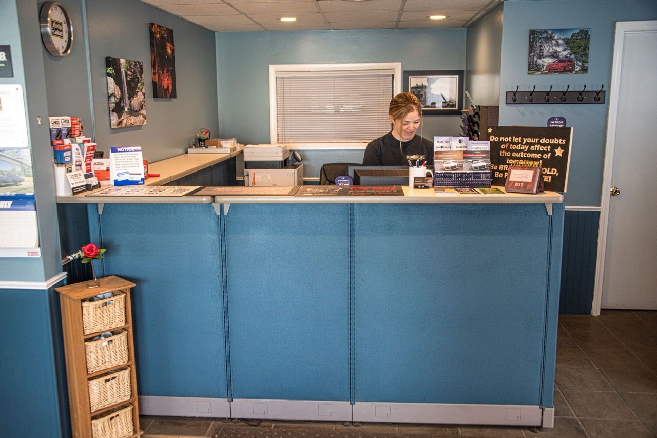 Receptionist at a desk in an auto repair shop. Blue walls, paperwork, and brochures | Jackson, MI