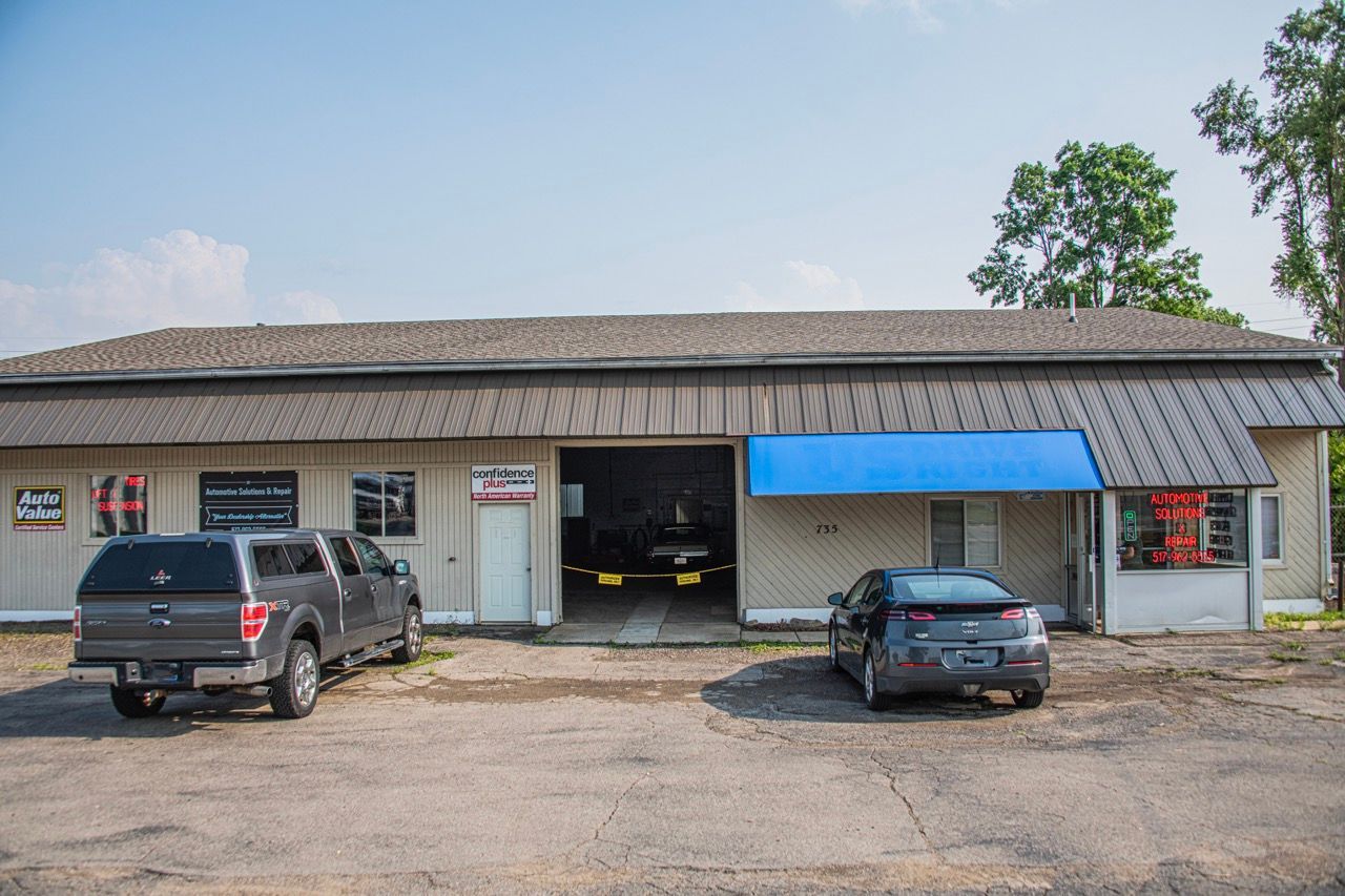 Auto repair shop with vehicles parked outside. Gray pickup and silver car. Open garage bay | Jackson, MI