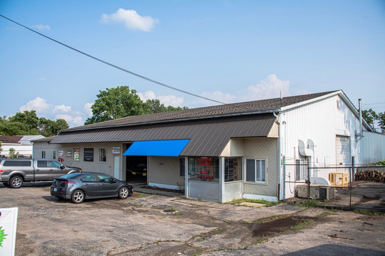 Exterior of a light-colored commercial building, blue awning, cars parked in front, clear sky | Jackson, MI