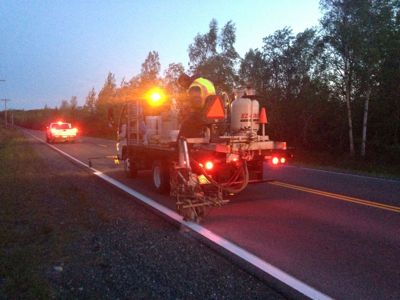 Road work vehicle with flashing lights on a highway at dusk, with another car in the distance