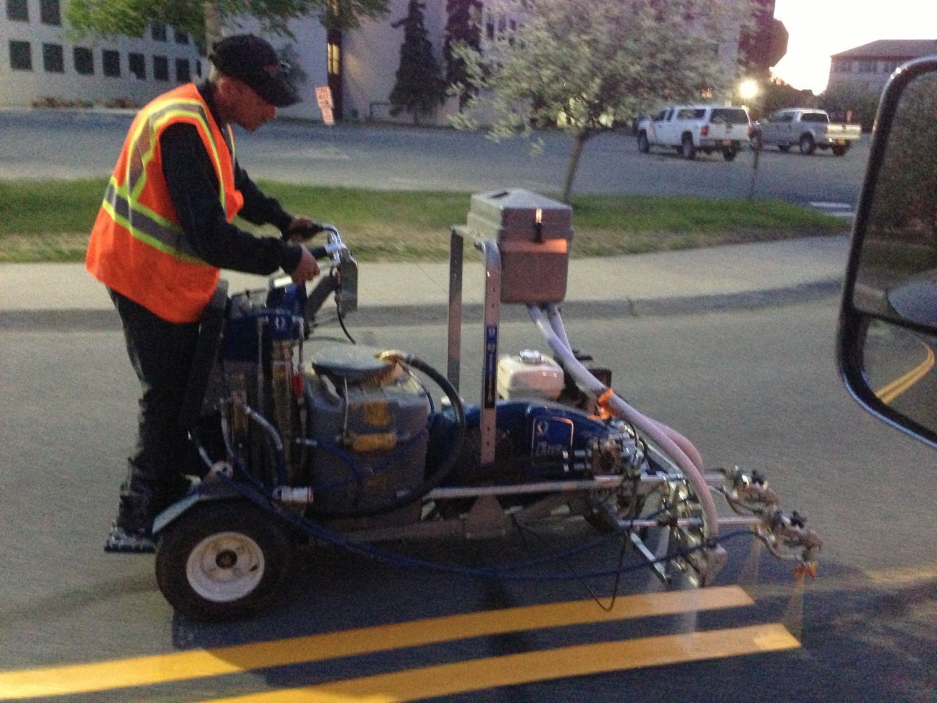 Worker in orange safety vest driving a small road-marking machine over yellow lane lines at dusk