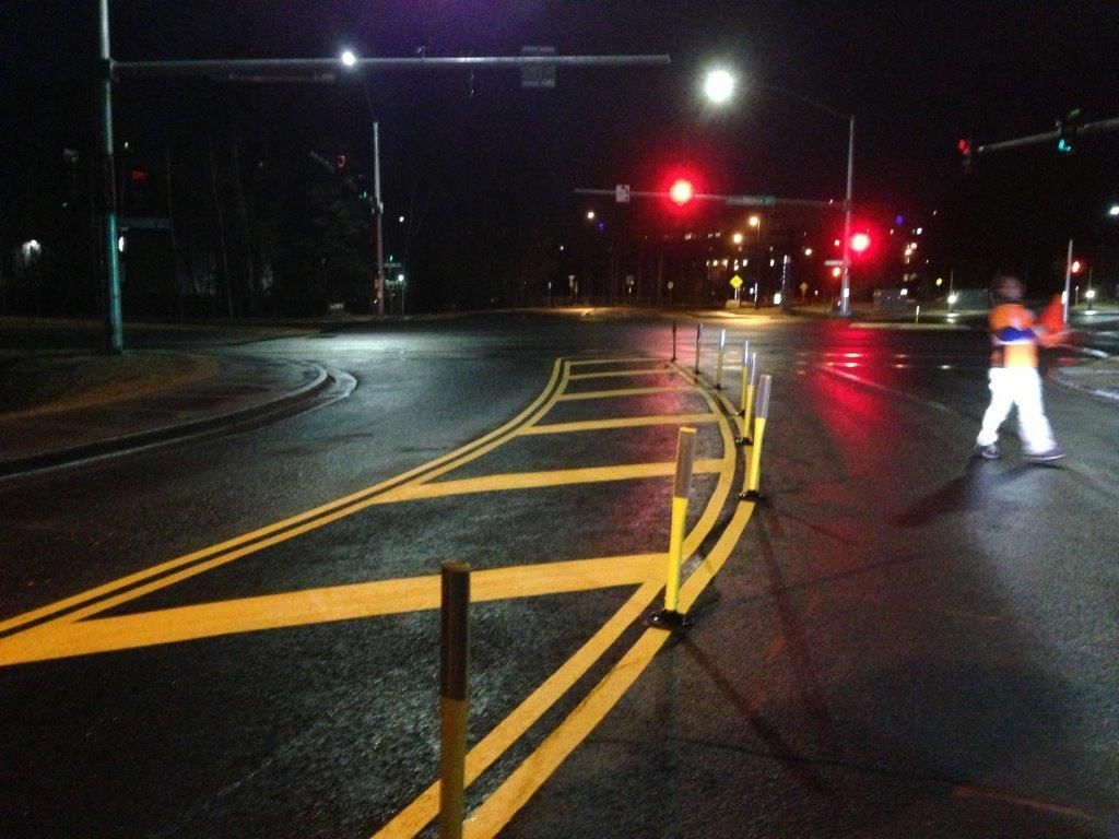 Nighttime roadwork with yellow barriers and a worker in reflective gear near red traffic lights.