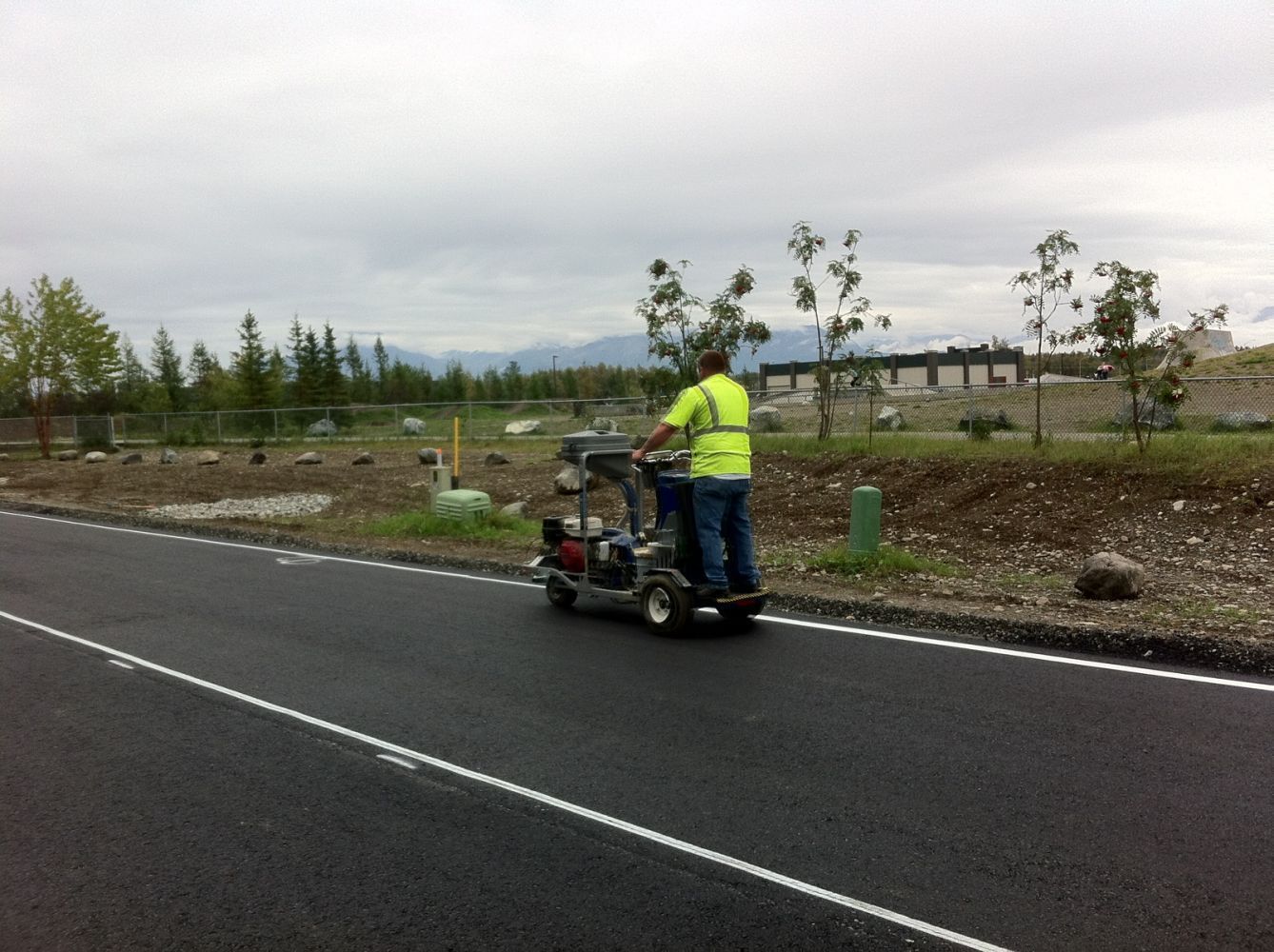 Worker in a neon jacket operates a road-marking machine on a wet roadside under cloudy skies