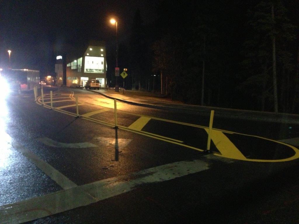 Nighttime street with a painted traffic island and yellow road markings near a lit building.