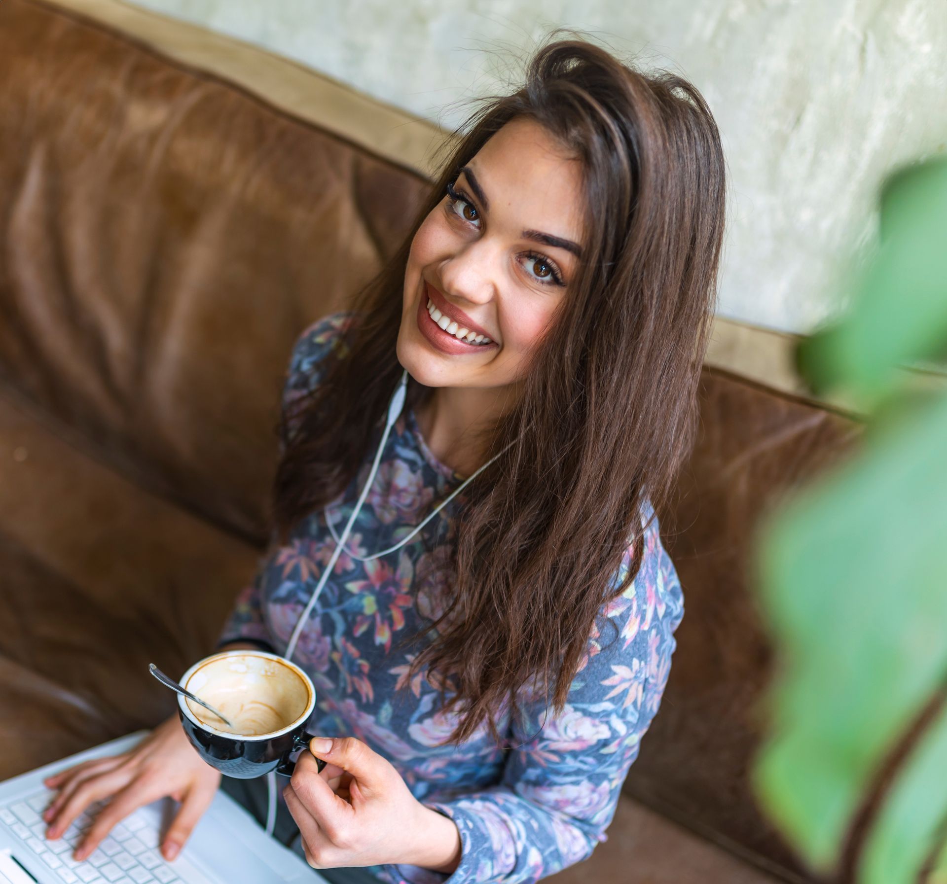 Woman holding coffee cup, smiling at the camera, wearing earbuds, sitting on a leather couch near a laptop.