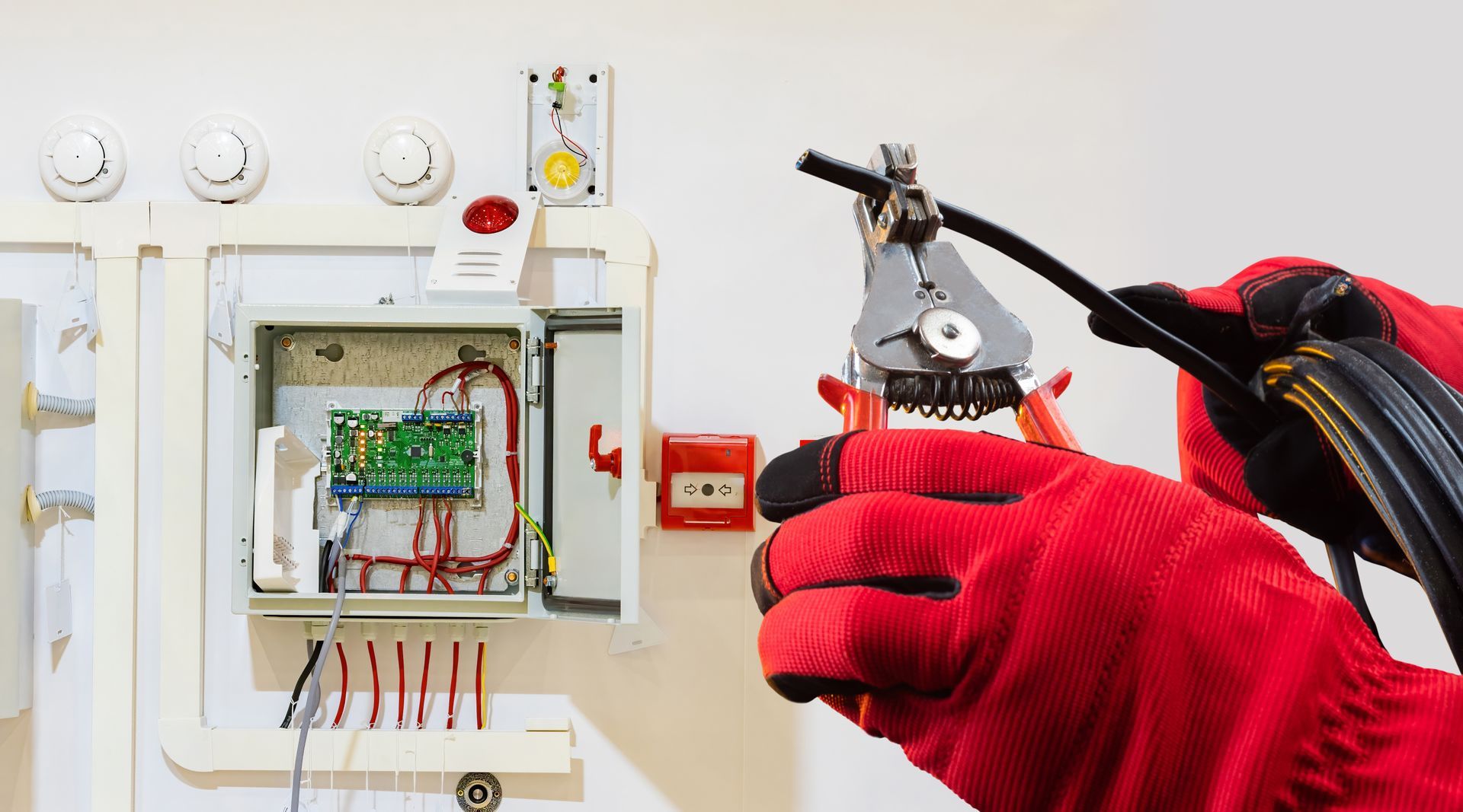 Gloved hands using pliers to work on exposed electrical wiring inside a wall panel