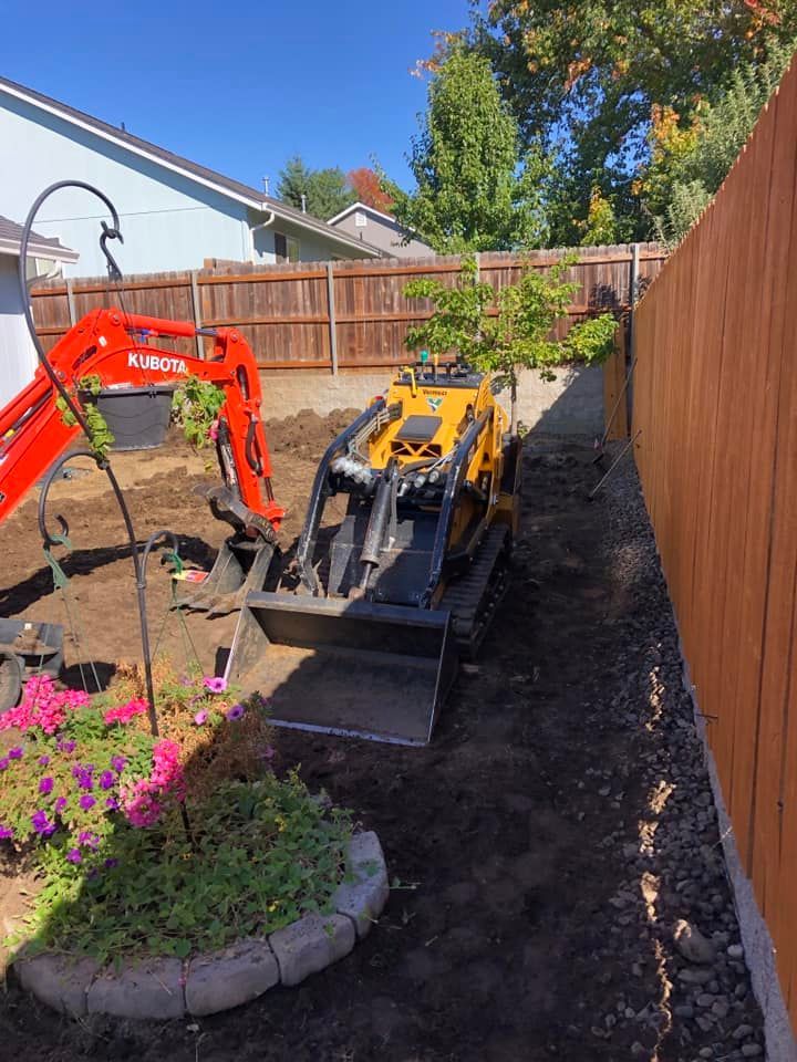Orange excavator and yellow mini skid steer working in a narrow backyard beside a wooden fence.