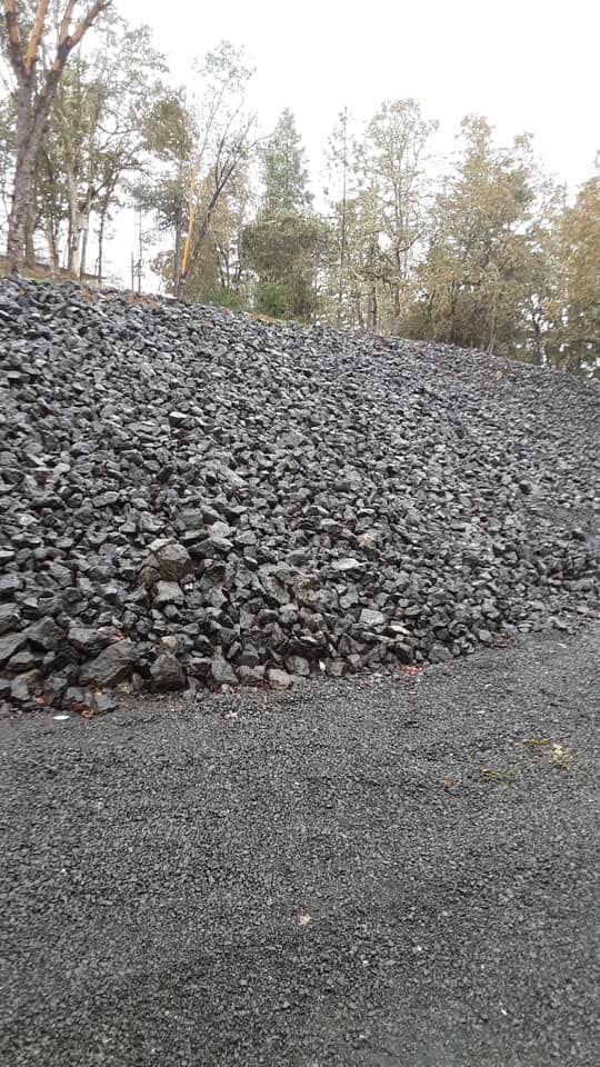 Large pile of dark gravel beside a dirt road with leafless trees in the background