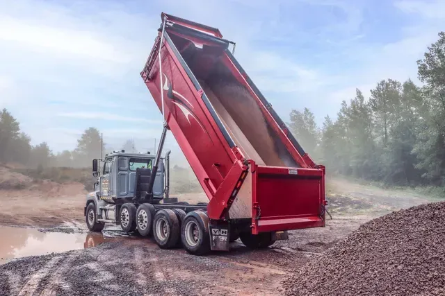 Red dump truck tipping gravel at a construction site, with dust rising and forest in the background