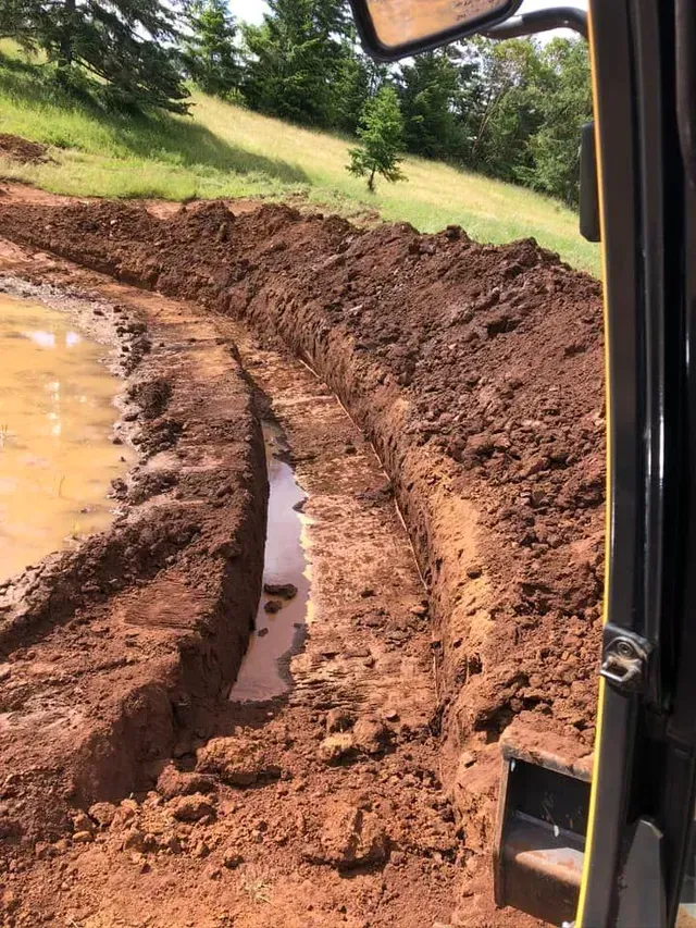 Mud-filled dirt road with deep tire ruts beside a waterlogged field, viewed from inside a vehicle