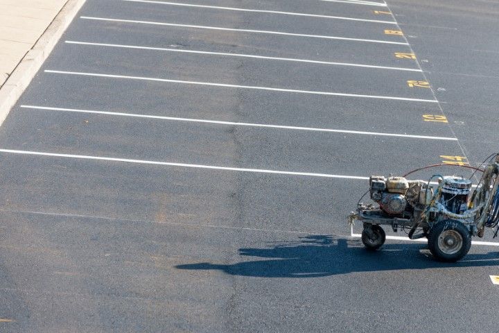 A machine paints white lines on an asphalt parking lot, with numbered spaces visible.